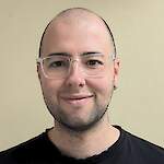 A smiling adult person with a shaved head, light stubble, and clear-framed glasses, wearing a black top against a plain light-colored background.