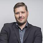 A man in a dark suit jacket poses confidently against a plain light-gray background.