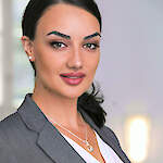 A woman with dark hair pulled back, light green/hazel eyes, and subtle makeup, wearing a gray blazer and white top.