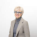 A woman smiles at the camera, wearing a grey suit and distinctive blue glasses