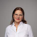 A woman with shoulder-length brown hair and patterned round glasses smiles softly.